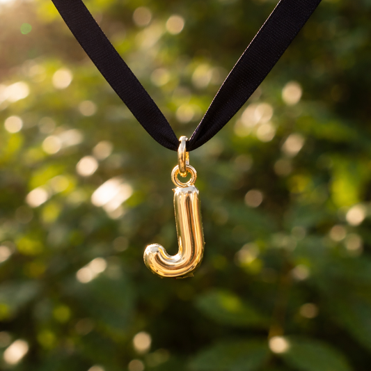 Gold 'J' pendant on a black ribbon against a blurred green background