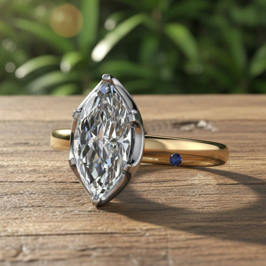 Gold ring with a large diamond and a smaller blue gemstone on a wooden surface with green foliage in the background.